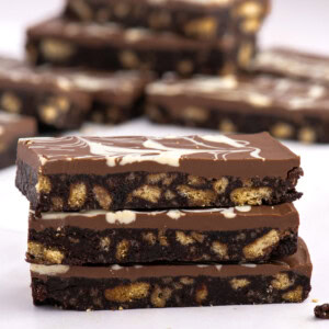 Stack of Scottish chocolate tiffin slices in focus, with a swirled chocolate topping and scattered biscuit pieces in the background.