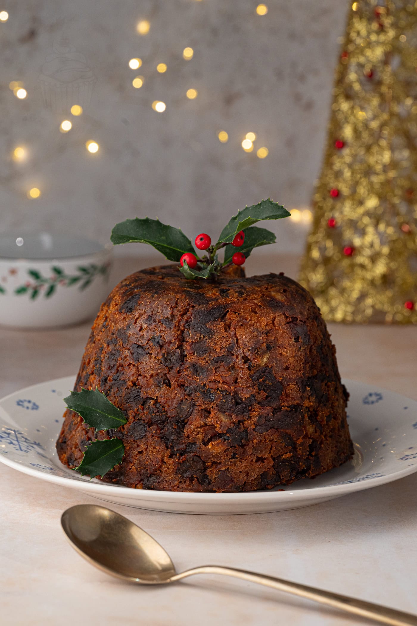 A traditional Christmas pudding sits on a decorative plate, topped with holly leaves and berries. In the background, festive lights twinkle softly while a gold decoration is partially visible. A spoon rests in the foreground, inviting you to savour this classic holiday delight.