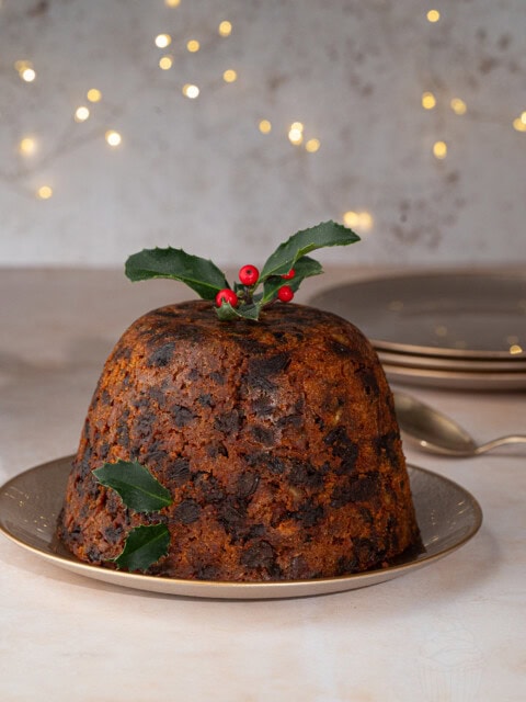 A traditional British Christmas pudding with a rich, dark texture sits on a plate. Its garnished with holly leaves and red berries. The background features soft, warm fairy lights and stacked plates.