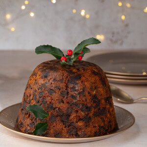 A traditional British Christmas pudding with a rich, dark texture sits on a plate. Its garnished with holly leaves and red berries. The background features soft, warm fairy lights and stacked plates.