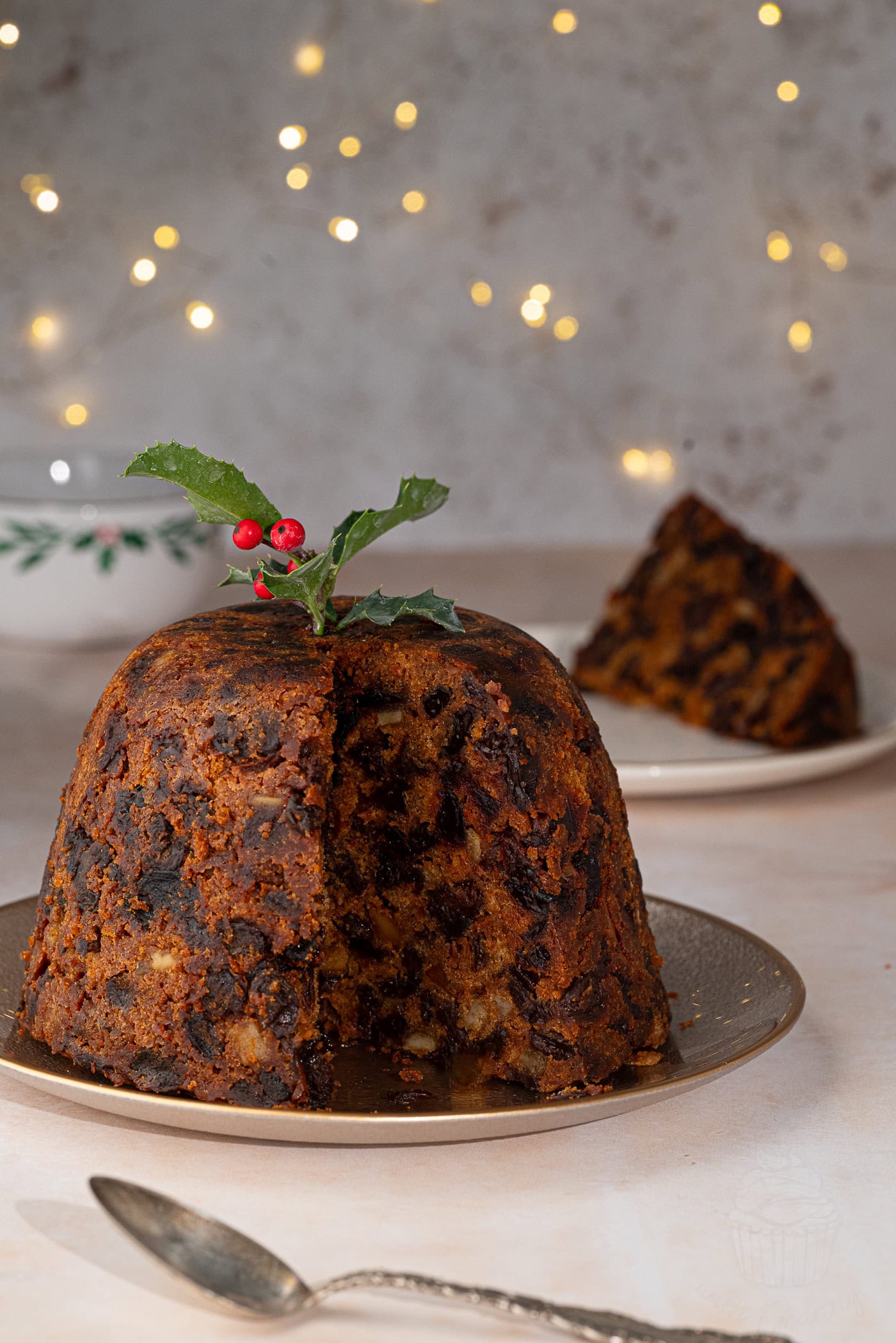 A traditional figgy pudding rests on a plate, adorned with a sprig of holly. A slice is served in the background, and festive lights softly illuminate the scene.