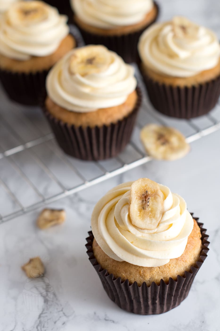 Banana Cupcakes recipe from Baking with Granny. Banana sponge cupcake, with a vanilla butter cream. A few more cupcakes sit behind the main one, with banana chips scattered around.