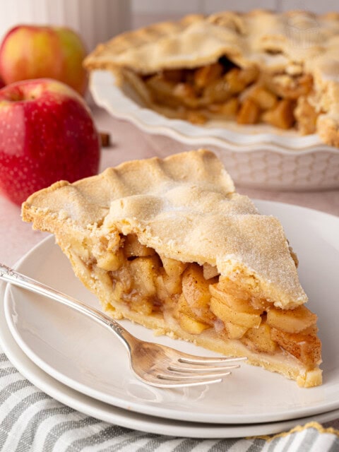Slice of apple pie on a plate with the full pie in the background. A classic apple pie UK recipe made with sweet, spiced apples.