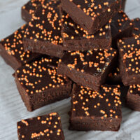 A stack of rich, sticky chocolate orange fudge topped with orange sprinkles, arranged neatly on parchment paper over a wooden surface. The vibrant sprinkles add a festive touch to the dark, fudgy goodness.