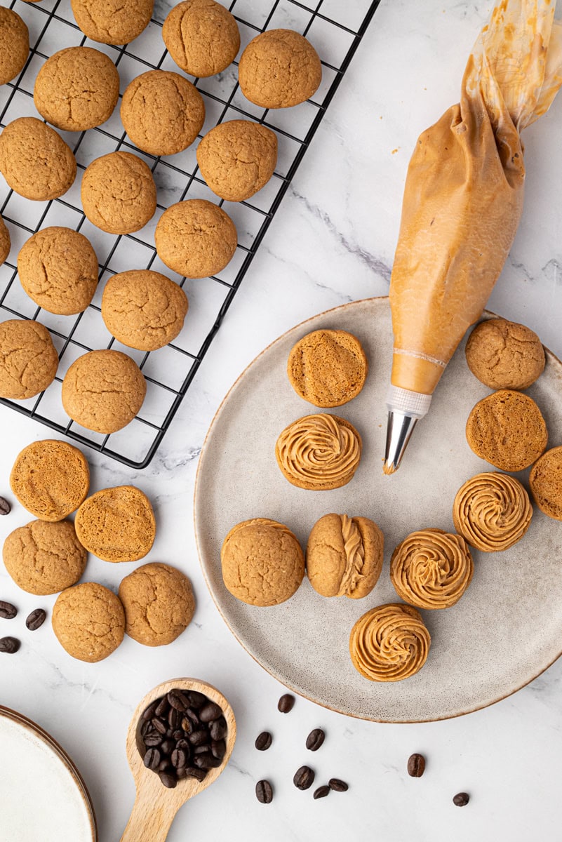 Overhead view of coffee kisses biscuits on a cooling rack with a piping bag of coffee buttercream and coffee beans.
