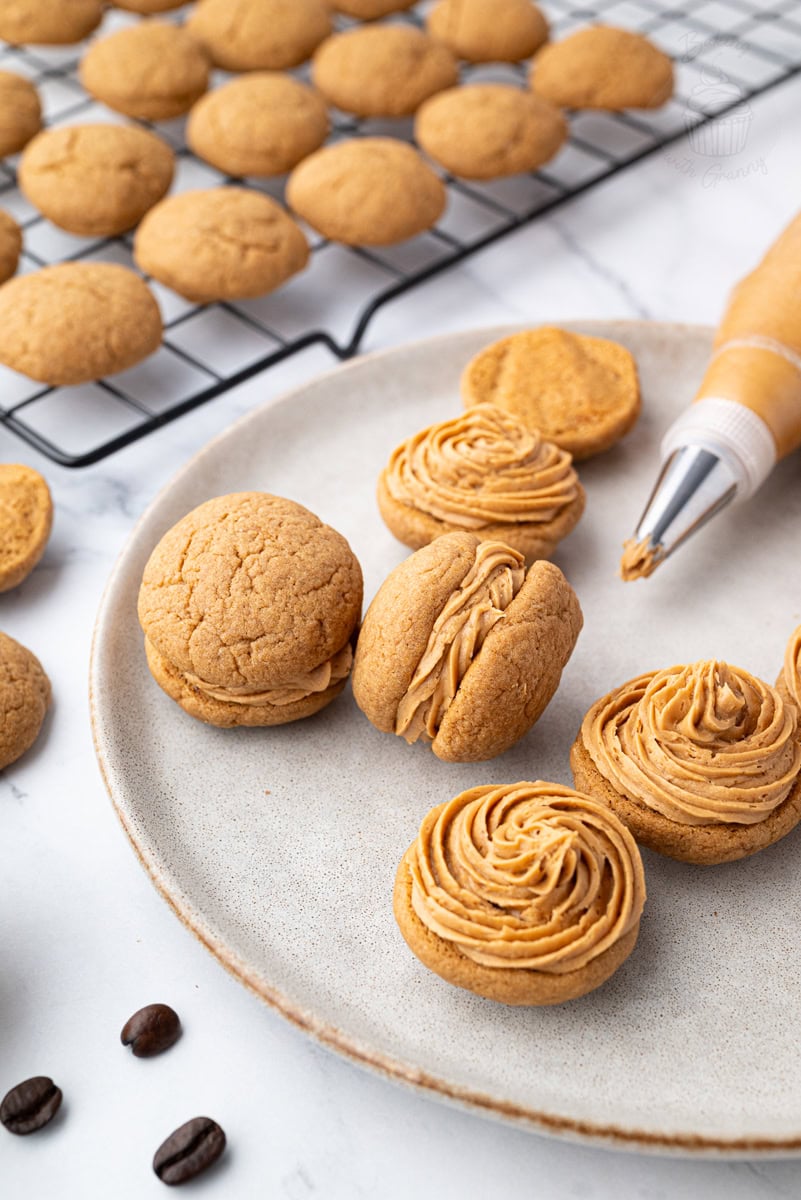 Coffee kisses biscuits being filled with piped coffee buttercream on a plate, with unfilled biscuits in the background.