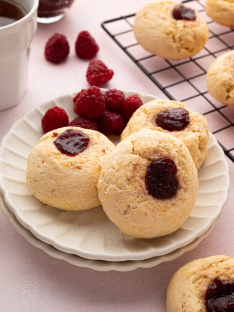 Three raspberry jam buns stacked on a plate with fresh raspberries and tea in the background.
