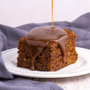 Warm sticky toffee pudding sponge with toffee sauce being poured over the top, dripping down the edges onto the plate.