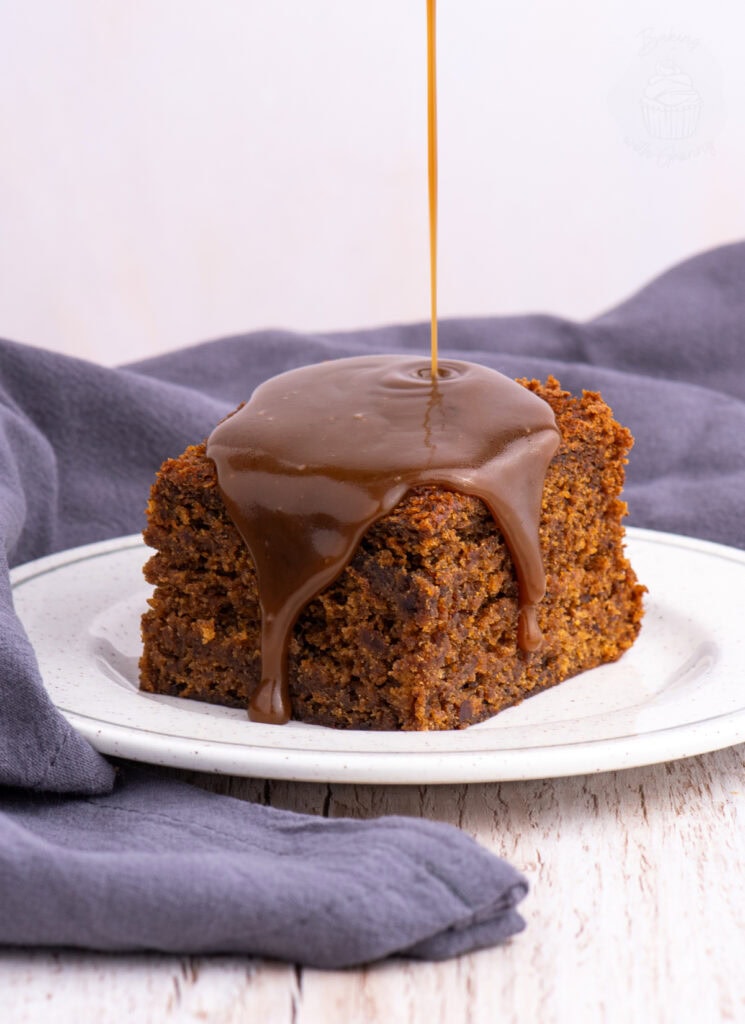 Warm sticky toffee pudding sponge with toffee sauce being poured over the top, dripping down the edges onto the plate.