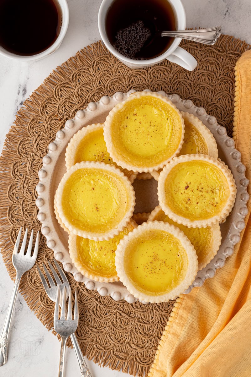 Overhead shot of custard tarts arranged on a plate with two mugs of tea and dessert forks alongside.