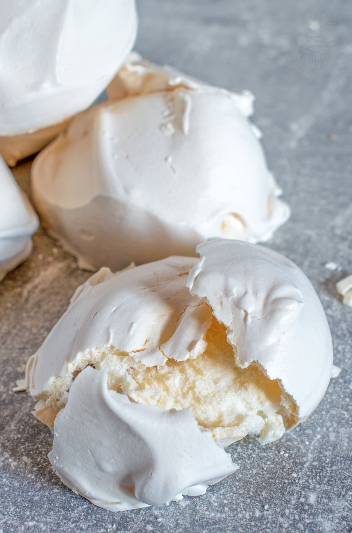Close-up of a broken homemade meringue showing its crisp shell and soft, chewy centre on a grey surface.