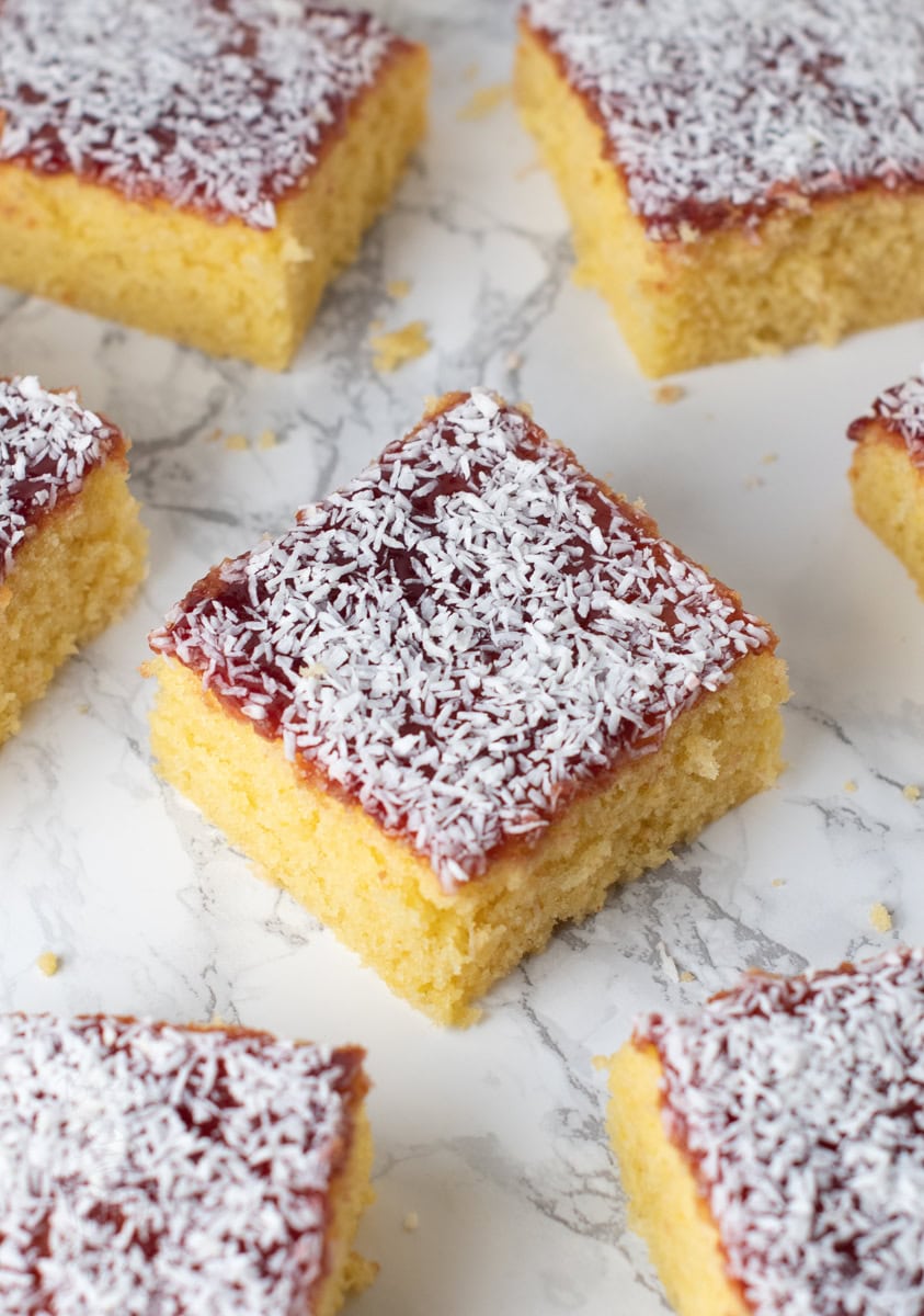 Overhead view of jam school cake squares on a marble surface, each topped with raspberry jam and desiccated coconut.