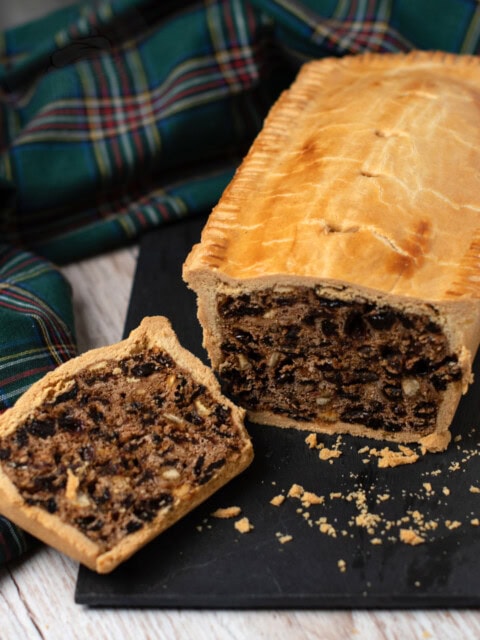 A Scottish black bun with a golden-brown crust rests on a black slate. One slice reveals a filling of dried fruit, nuts and raisins. A green and blue tartan cloth is in the background on a wooden surface.
