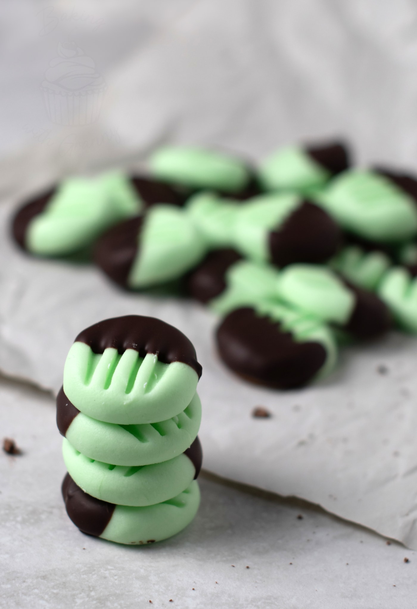 A stack of mint green peppermint creams, which are partly dipped in chocolate, with more patties scattered on parchment paper in the background.