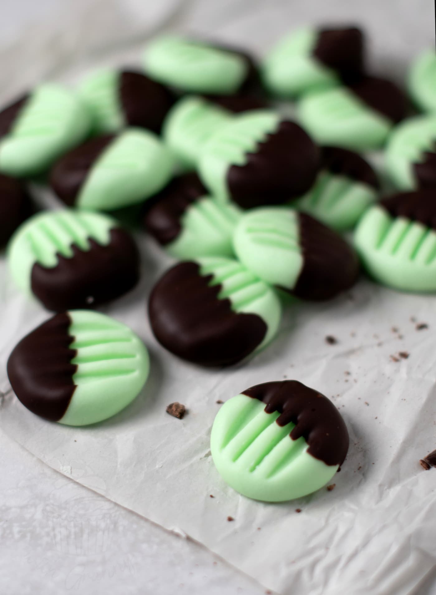 Mint chocolate peppermint creams with a green base and dark chocolate-dipped tops. Scattered on crumpled parchment paper, these peppermint fondant creams are adorned with a fork imprint pattern on the tops.