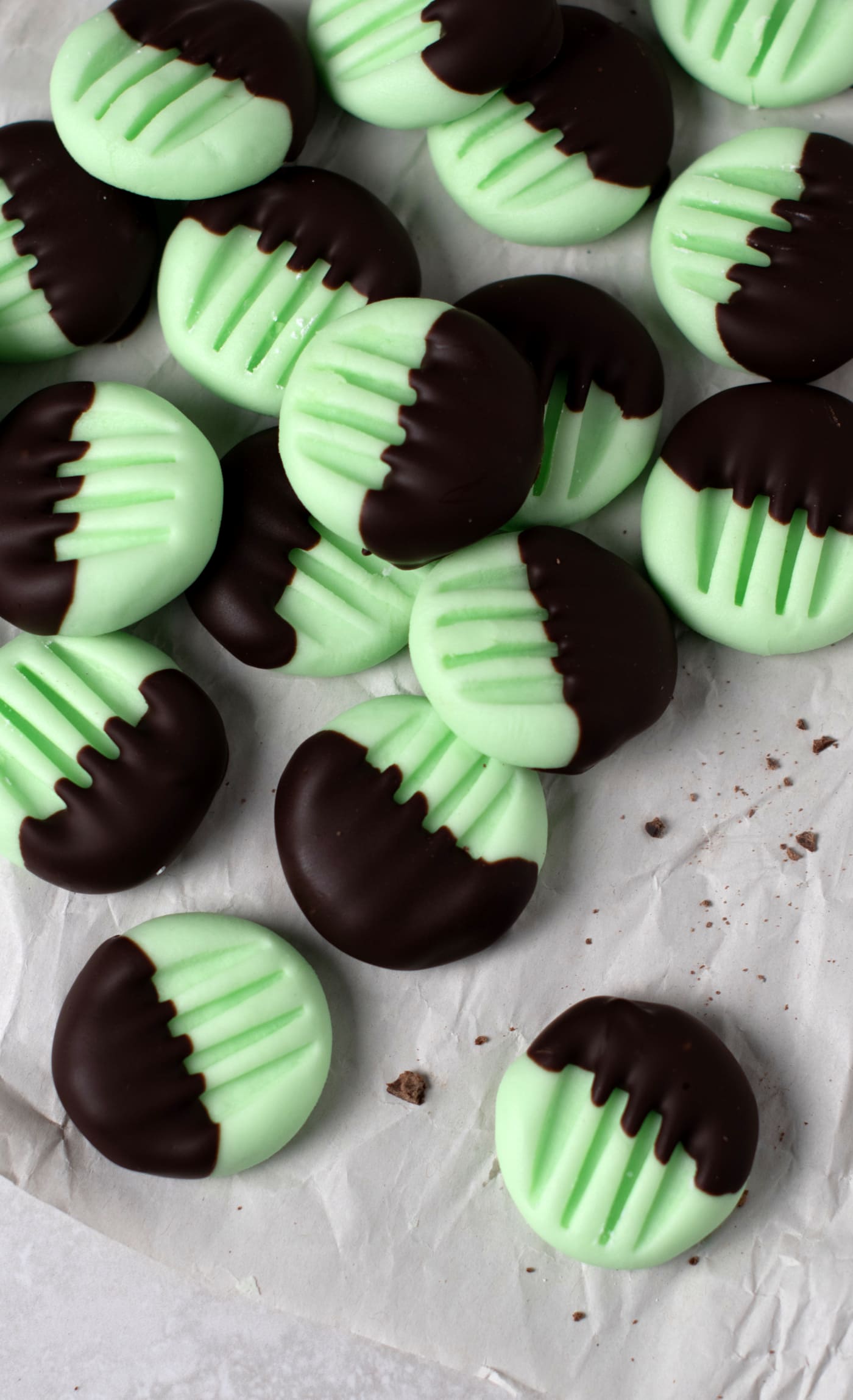 A pile of round, green peppermint creams half-dipped in dark chocolate are scattered on crumpled white paper. These traditional peppermint creams have a grooved pattern on their tops. Chocolate crumbs speckle the paper