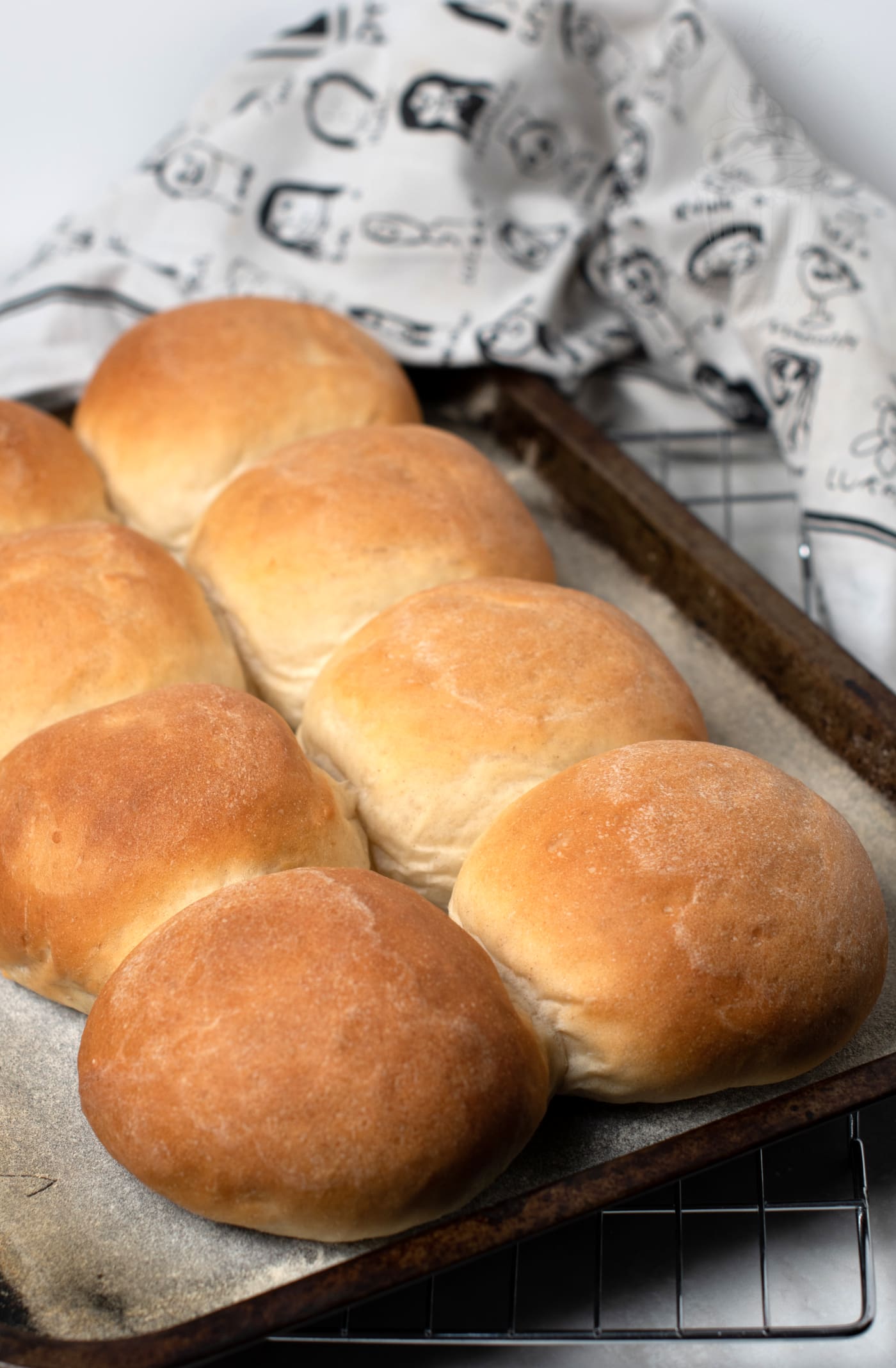 A tray of eight freshly baked morning rolls rests on parchment paper. The rolls are golden brown, enticing anyone trying out this recipe, while a patterned cloth is partially visible in the background.