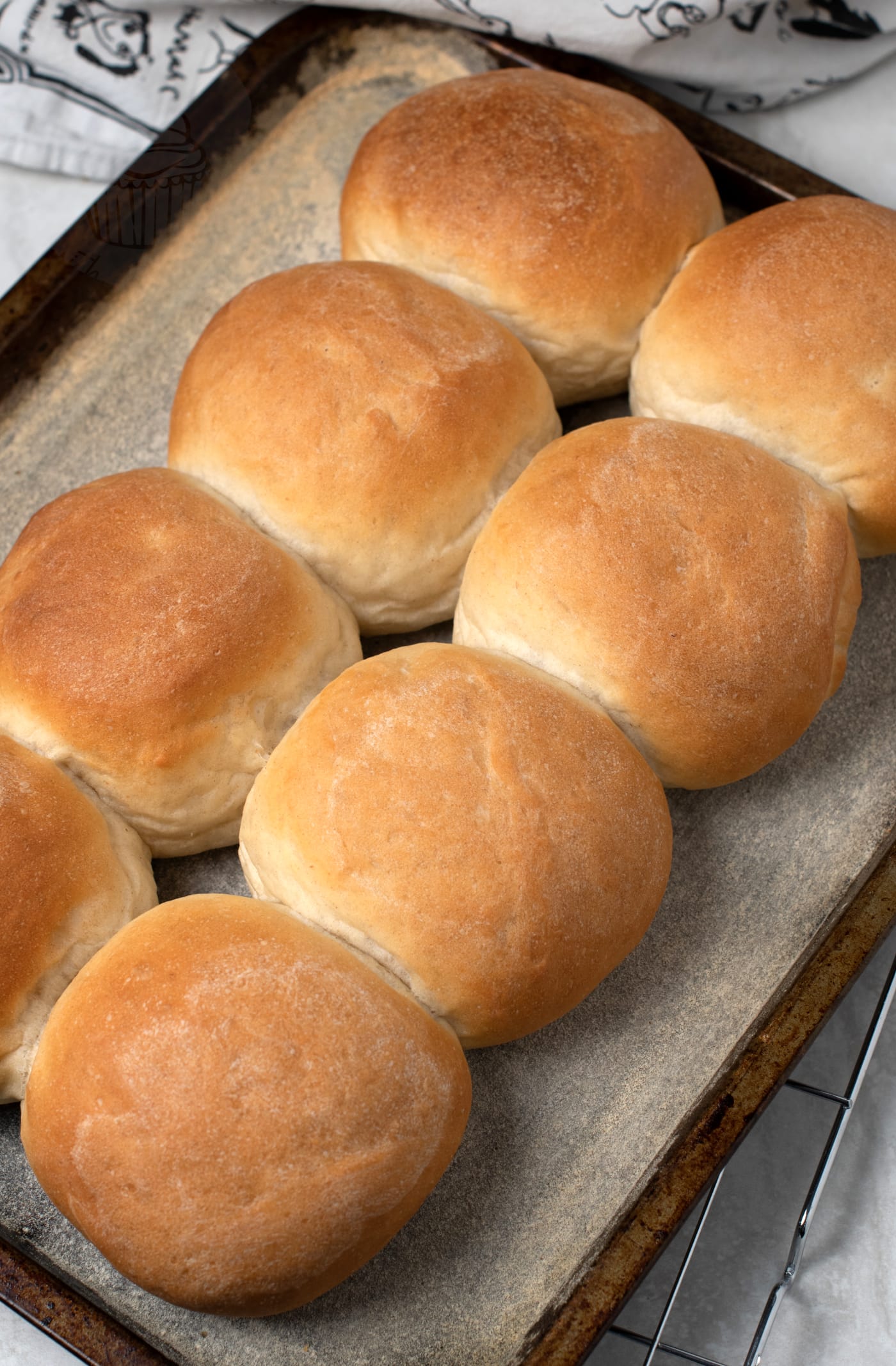 A tray of eight freshly baked golden brown Scotch morning rolls is arranged in two rows of four on parchment paper. The rolls have a soft, smooth surface and appear fluffy. The tray rests on a cooling rack, with a patterned cloth nearby.