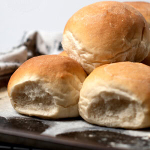 A close-up of freshly baked Scottish morning rolls on a baking tray. The rolls are golden brown with a slightly cracked, crusty surface. A white cloth is partially visible in the background, hinting at their homemade recipe origin.