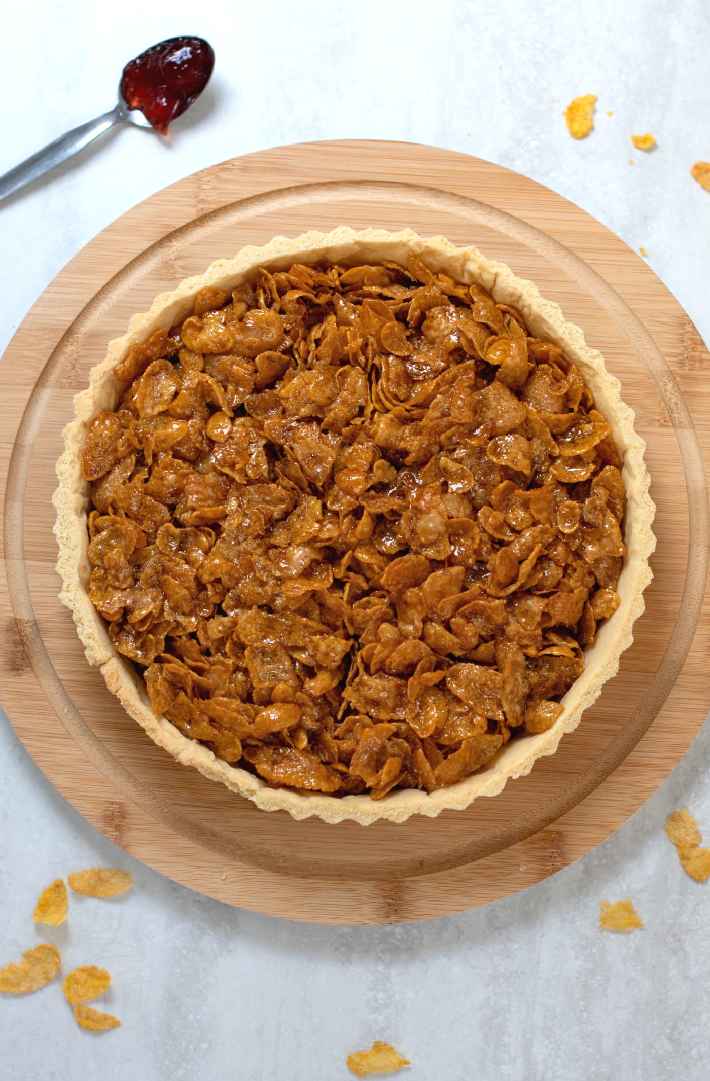 Cornflake tart from an aerial view. The crunch topped tart sits on a round wooden board, with a spoonful of jam to the side, and a scattering of extra cornflakes around.