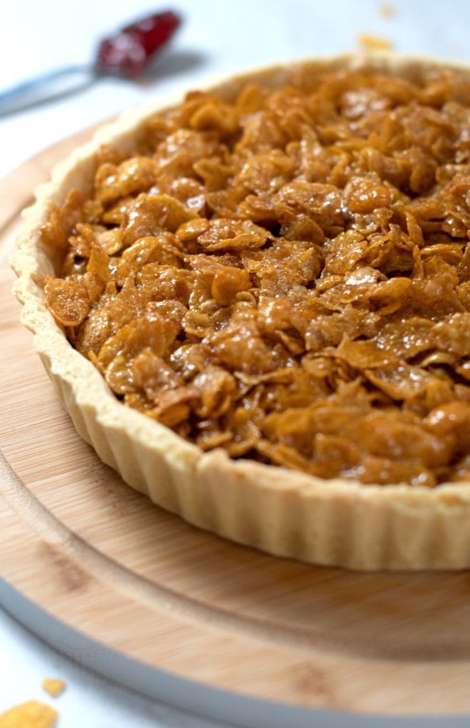 Close up of a golden cornflake tart, on a round wooden board, with a spoonful of jam in the background.