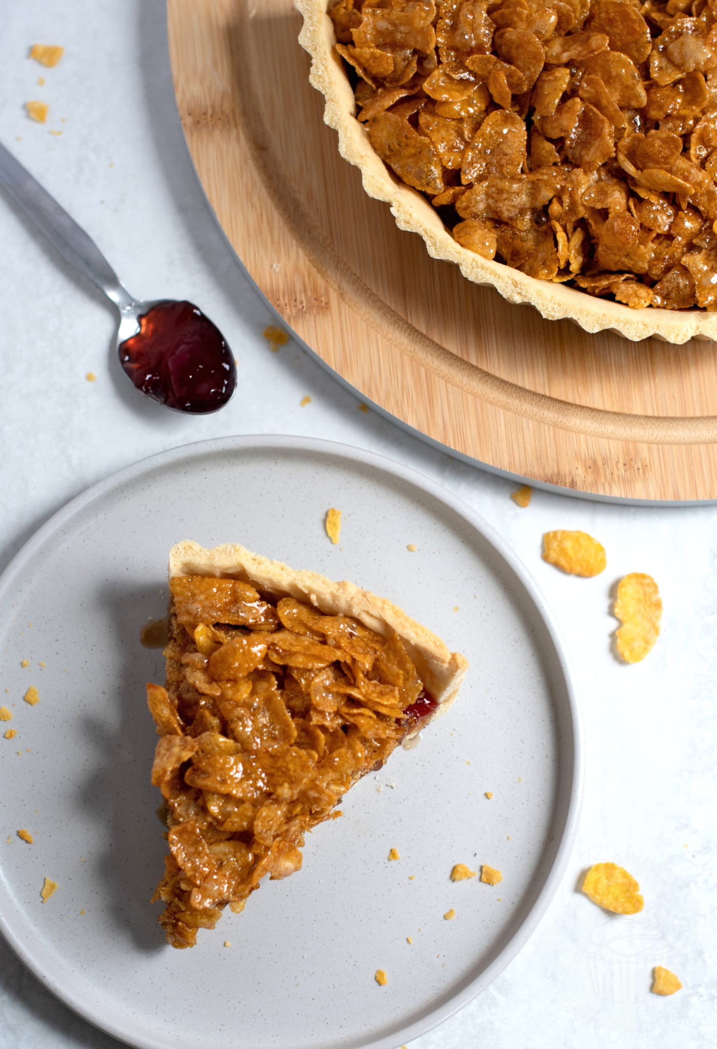 Overhead view of a slice of cornflake tart on a grey plate, with the rest of the tart on a circular wooden board to the side. A spoonful of jam and broken cornflakes are scattered around on the white work surface.