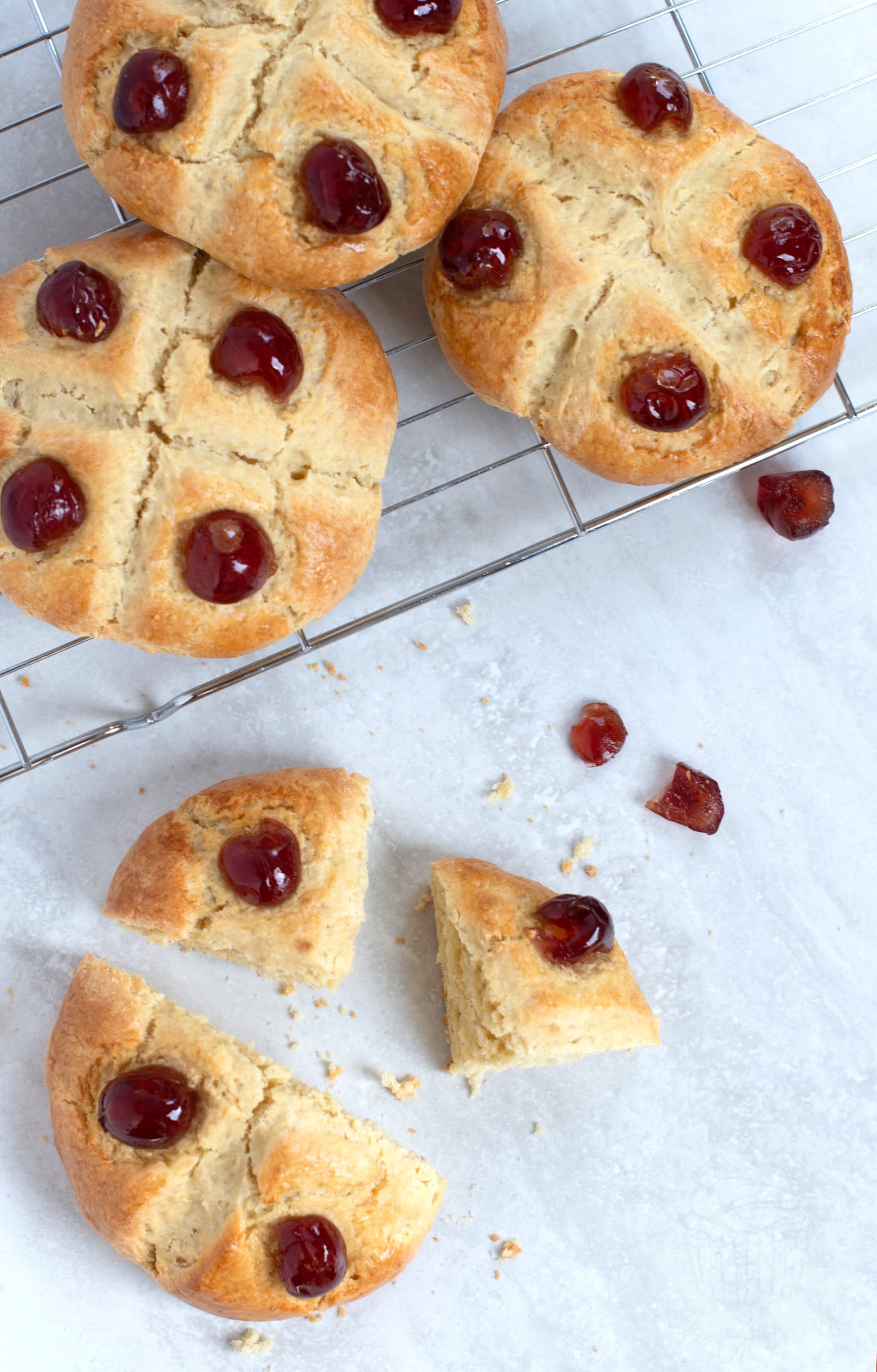Victoria scones on a wire rack. One of scones in cut into quarter triangles, ready to serve. Small pieces of excess glace cherries are scattered around.