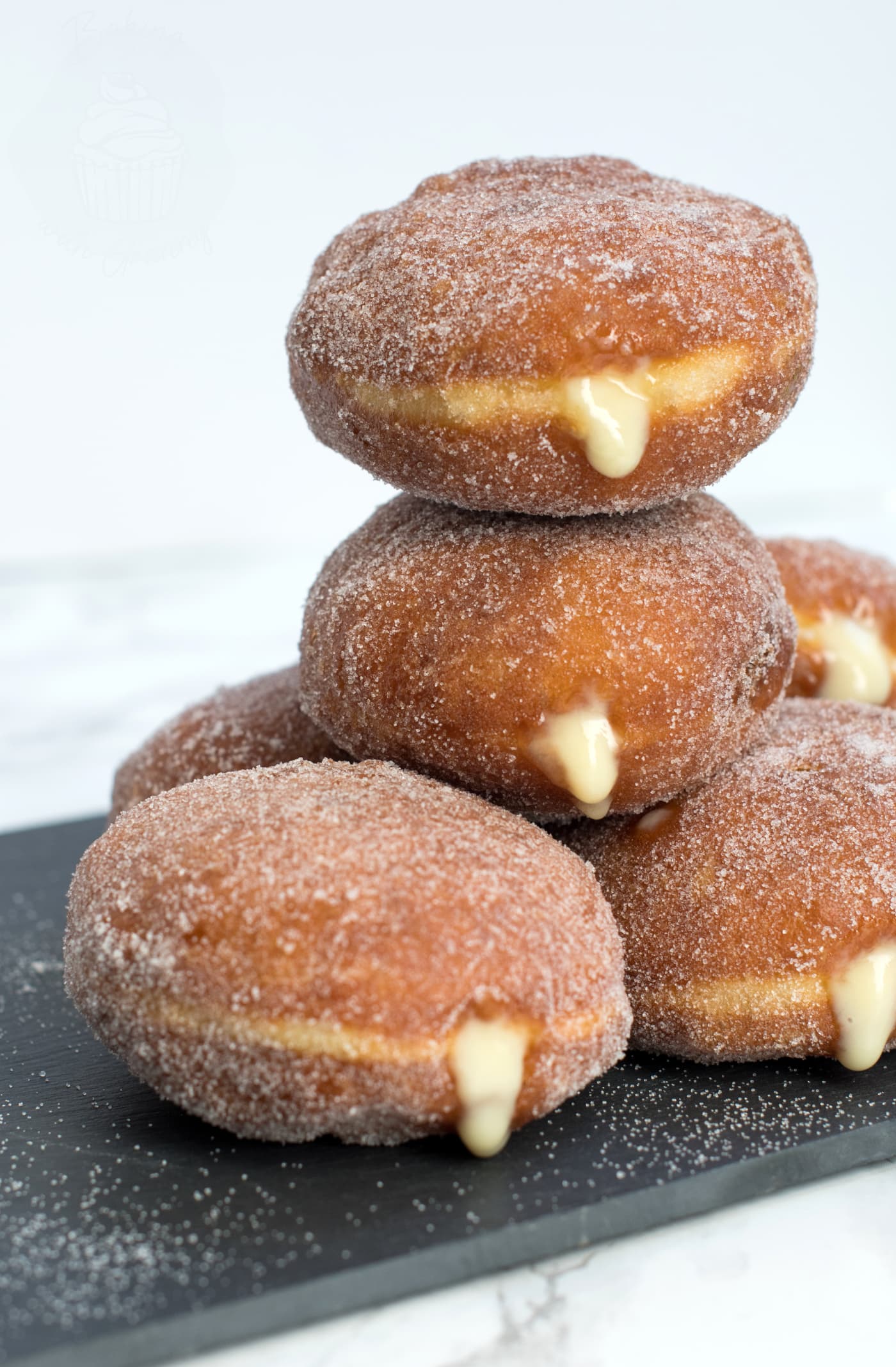 Stack of custard doughnuts on a black slate chopping board. The custard can be seen oozing from the sides of the sugar coated doughnuts.