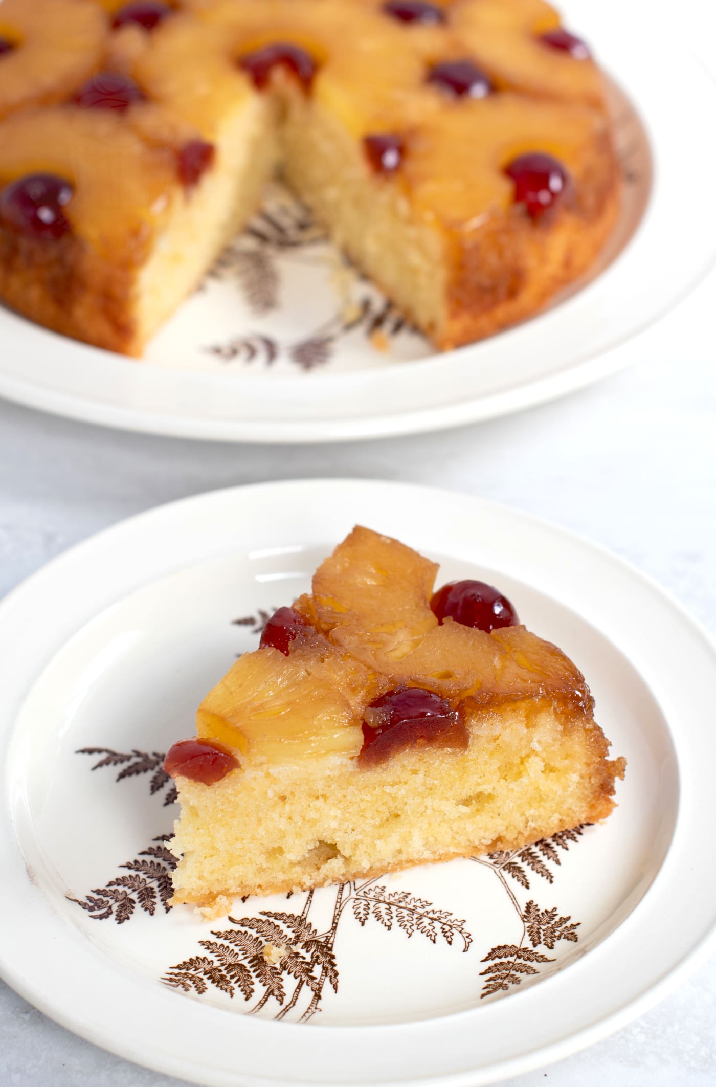 Slice of Pineapple Upside Down Cake, with the rest of the cake in the background. The inside of the sponge cake is visible, and a light golden in comparison to the pineapple rings on top.