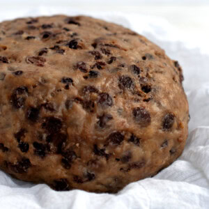 A round, dense traditional Scottish clootie dumpling, filled with visible raisins and currants, rests on a white cloth. The dumplings textured surface reflects its rich, moist composition.