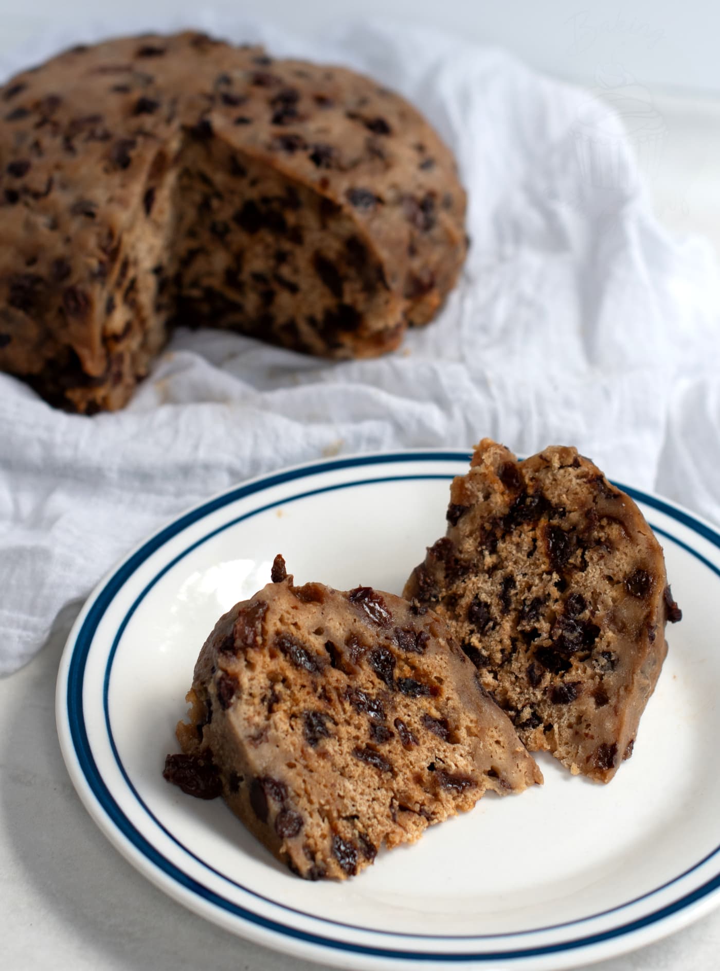 A sliced clootie dumpling with dark fruit pieces rests on a white plate with a blue rim. The traditional pudding, partially cut, sits invitingly on a white linen cloth (also known as a "cloot") in the background.