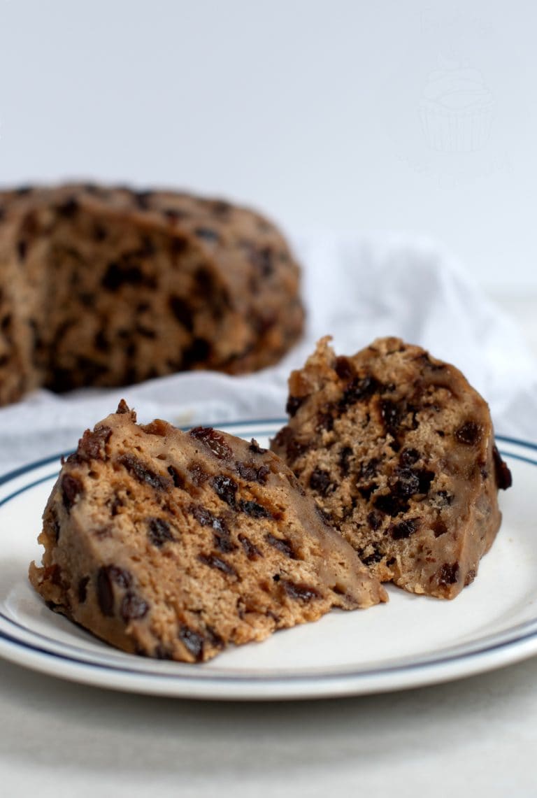 Two slices of traditional clootie dumpling with raisins and spices rest on a white plate with a blue rim, made from a classic Scottish clootie dumpling recipe. A larger portion of the dumpling is blurred in the background on a white surface.