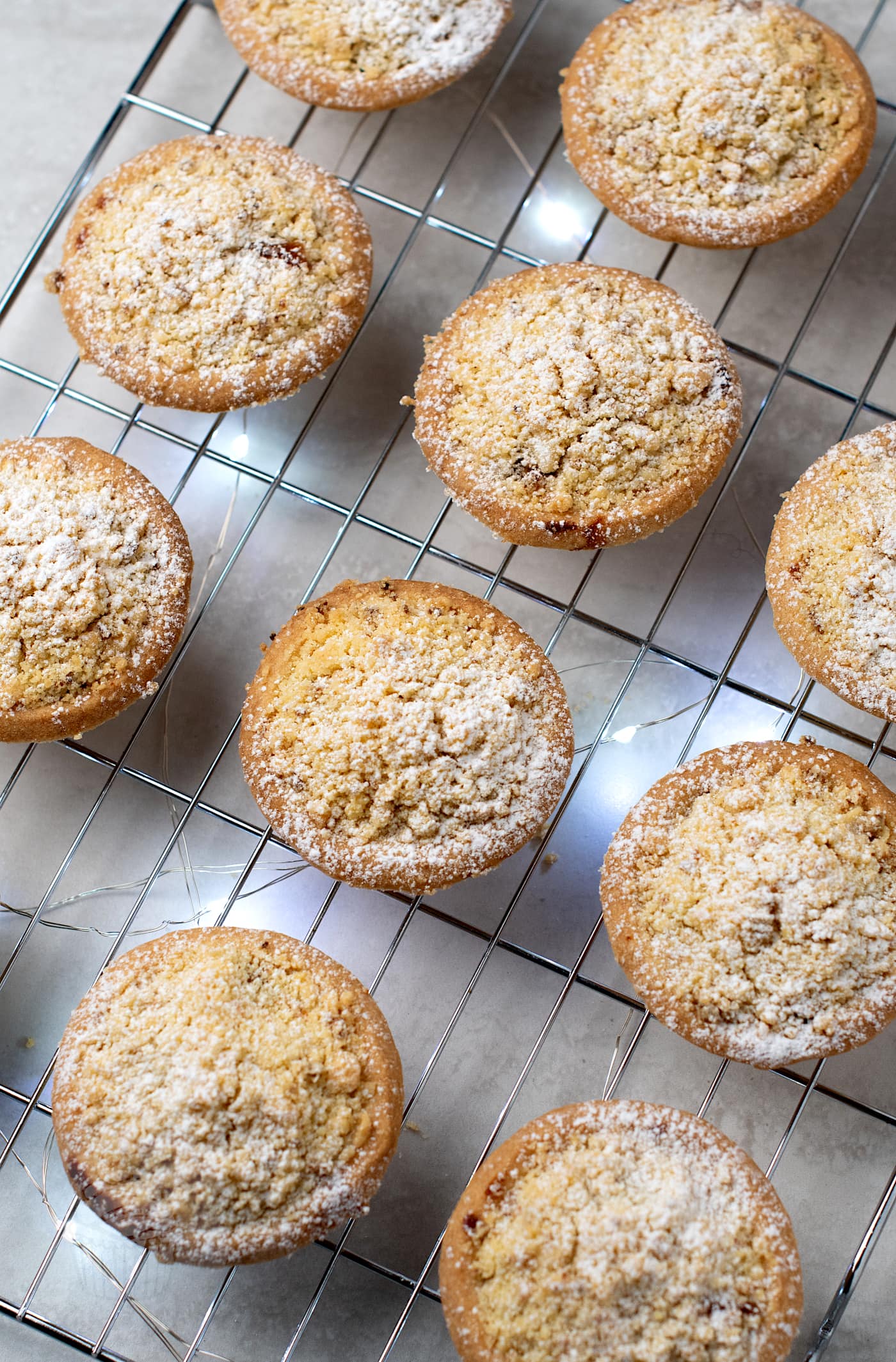 Ten freshly baked mince pies with a crumble top are cooling on a wire rack. They are evenly spaced on a light-coloured countertop, and the festive mince pies have a golden-brown hue.