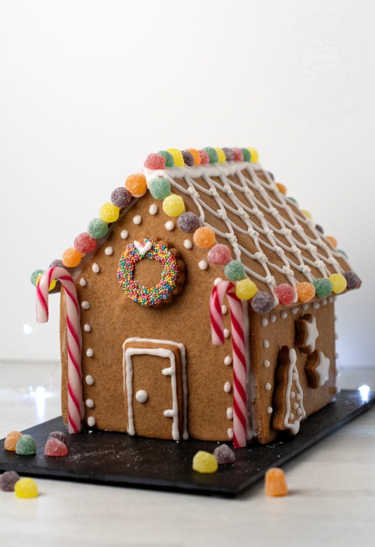 A gingerbread house crafted from a delightful UK recipe is adorned with colourful jelly tots, striped candy canes, and white icing patterns. A wreath made of sprinkles graces the front. The house rests on a black slate surrounded by scattered sweeties.