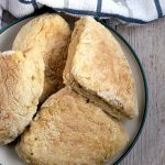 A plate of freshly baked, triangular shaped traditional Irish soda farls, sits on a wooden table. A striped white and blue kitchen towel is placed nearby.