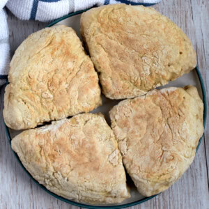 Show thumbnail preview A plate with four freshly baked, round traditional Irish soda farls, placed on a wooden surface. A striped kitchen towel is visible in the background, hinting at a cherished soda farls recipe just waiting to be recreated.