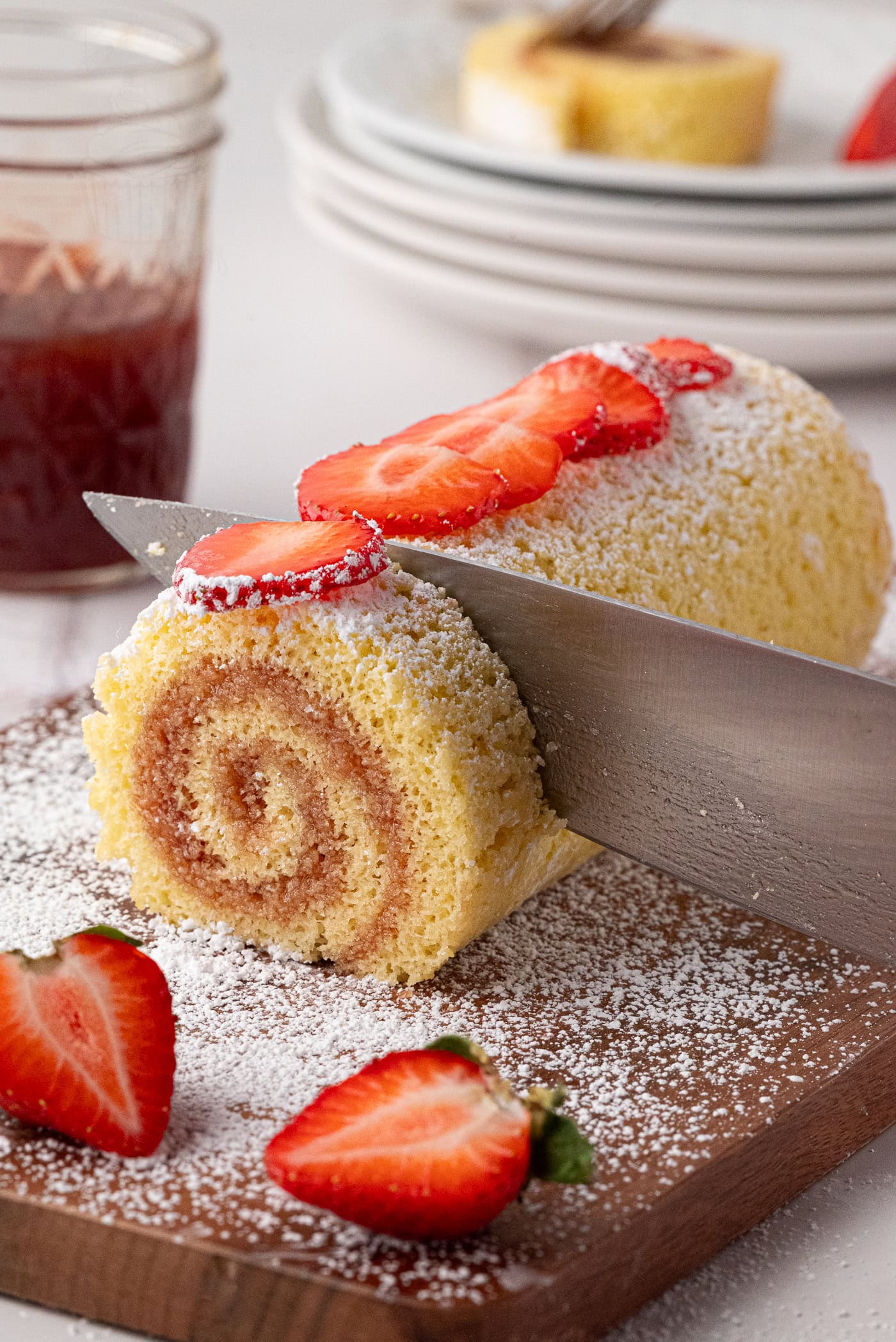 Swiss Roll A knife slices into a icing sugar-dusted Swiss roll, topped with fresh strawberry slices. More strawberries are scattered on the wooden cutting board, and a jar of jam along with stacked plates are in the background, echoing the timeless charm of a classic UK recipe.
