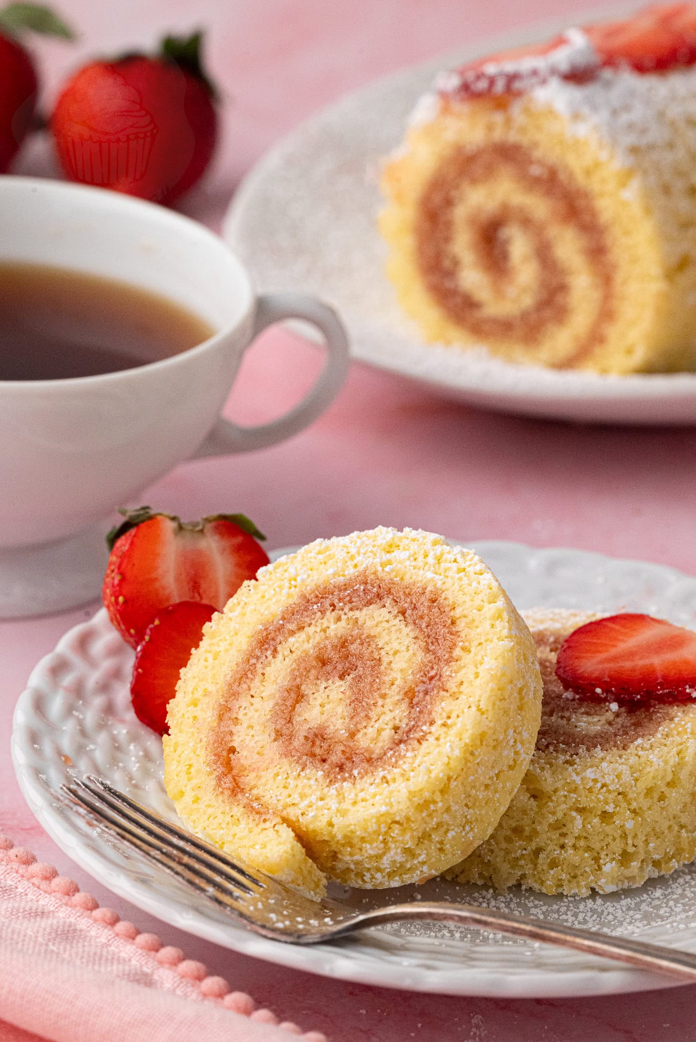 A close-up of a delicious Swiss Roll cake slice, made from an easy Swiss Roll Recipe, sits on a white plate, garnished with strawberry slices. A fork rests beside it. In the background, a cup of tea and another slice of cake on a pink surface are visible.