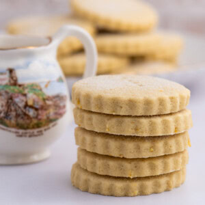 A stack of round, scalloped-edge traditional Scottish Tantallon Cake biscuits sits next to a decorative jug with a landscape design of North Berwick. More cookies are blurred in the background, all on a light surface, evoking the charm of an age-old recipe.