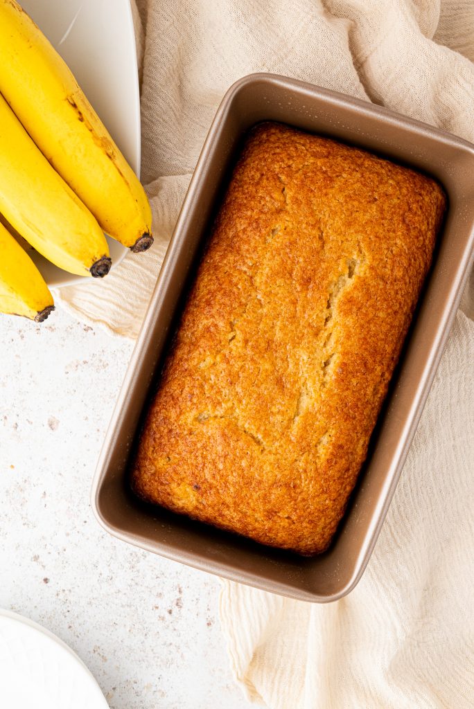 A freshly baked banana loaf, made from a beloved family recipe, sits invitingly in a baking pan on the light countertop. Nearby, three ripe bananas rest on a white plate, while a beige cloth peeks out from beneath the pan.