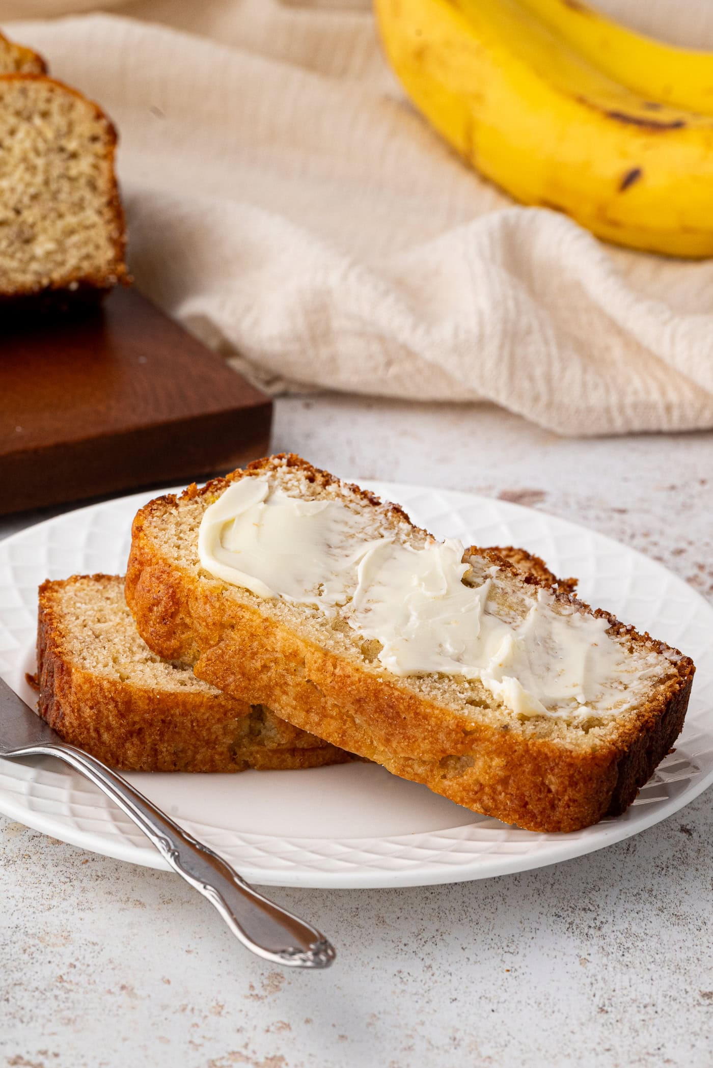 Two slices of banana bread, illustrating a delectable banana bread recipe, sit on a white plate with butter spread on top. Nearby, a fork awaits indulgence. In the background, a whole banana and fresh loaf rest on a wooden board atop a beige cloth.