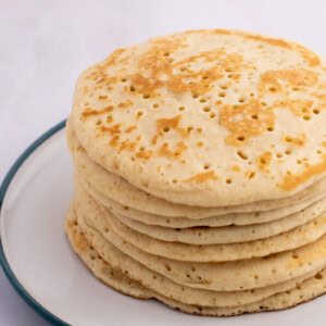 A stack of eight bubbly Scottish crumpets with a light golden-brown colour, features small holes on the surface and sits on a white plate with a green rim. The background is a soft, blurred white.