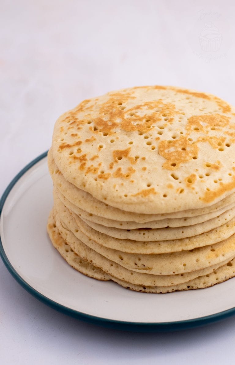 A stack of eight bubbly Scottish crumpets with a light golden-brown colour, features small holes on the surface and sits on a white plate with a green rim. The background is a soft, blurred white.