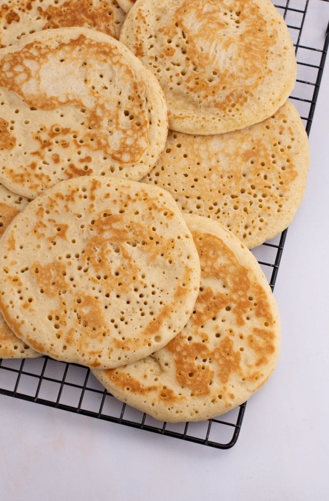Crumpets-042-2 Several round crumpets with golden-brown tops and characteristic holes, reminiscent of a traditional Scottish crumpet recipe, are cooling on a black wire rack placed on a white surface.