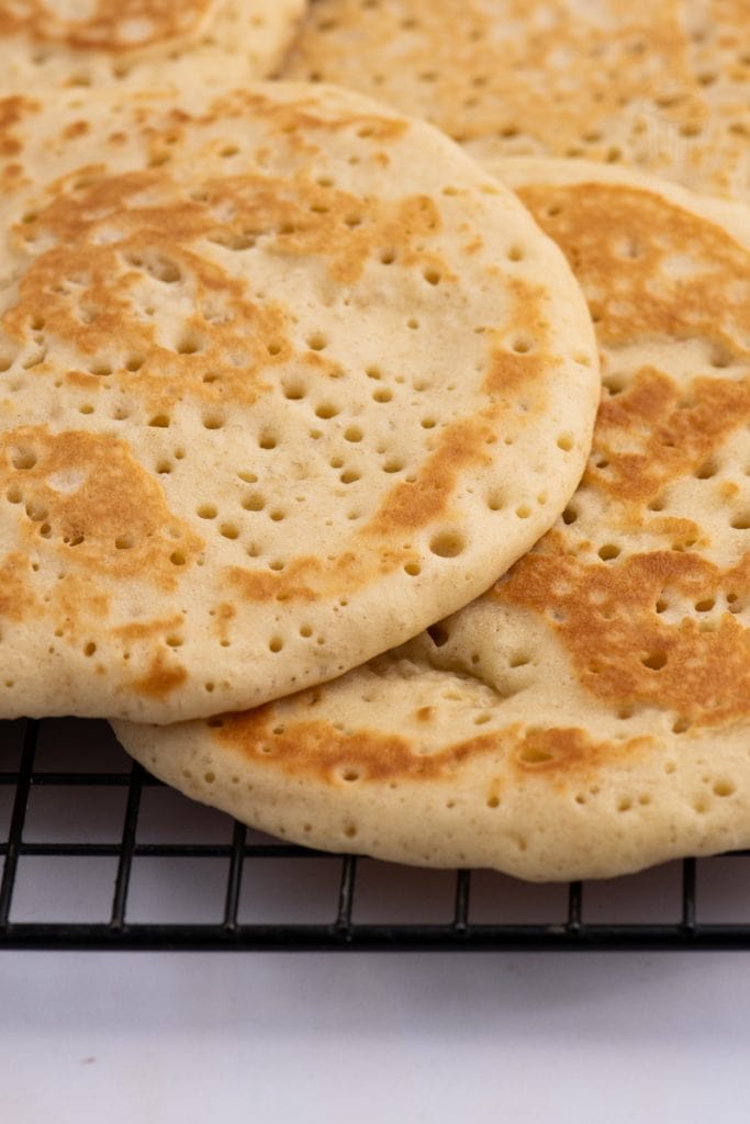 Close-up of a stack of classic Scottish crumpets with a golden-brown surface, featuring characteristic bubbles and light texture, resting on a black wire cooling rack.