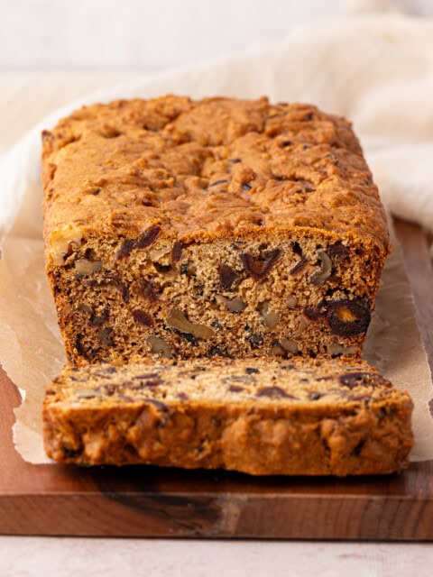 A loaf of date and walnut cake sits on a wooden board, with one slice cut and placed in front. The cake appears rich and dense, filled with nuts and dried fruits. Its resting on a piece of parchment paper, with a light-coloured cloth gently draped in the background.