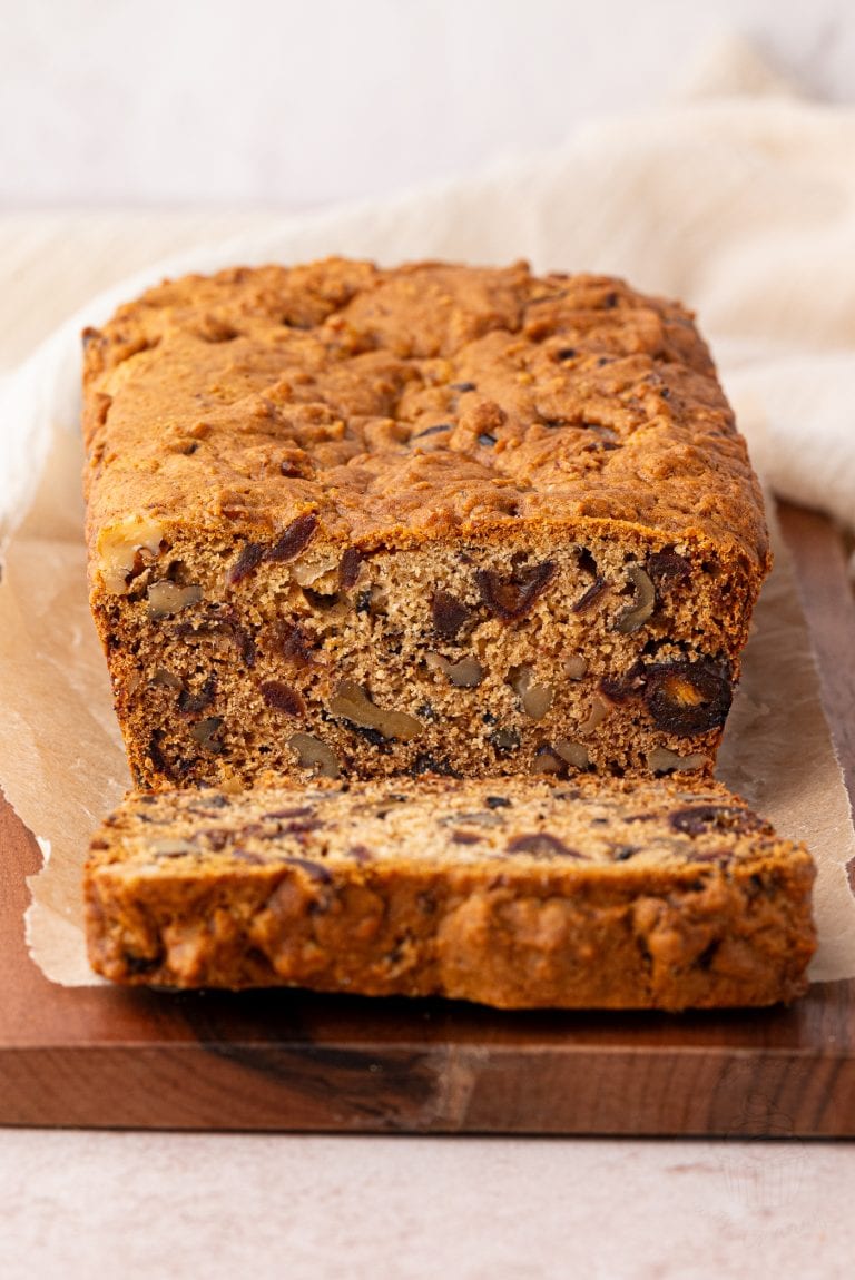 A loaf of date and walnut cake sits on a wooden board, with one slice cut and placed in front. The cake appears rich and dense, filled with nuts and dried fruits. Its resting on a piece of parchment paper, with a light-coloured cloth gently draped in the background.