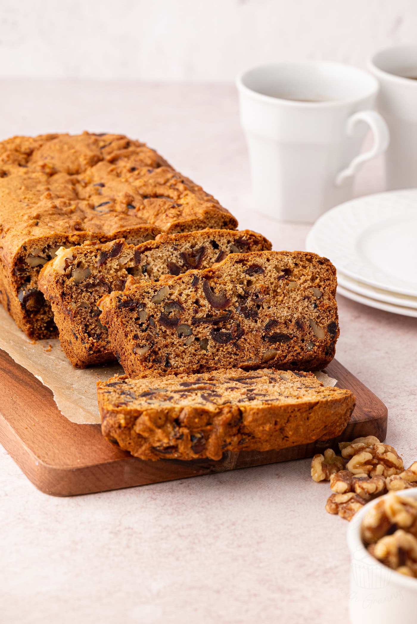 Sliced walnut and date loaf on a wooden cutting board with two coffee cups and saucers in the background. A small bowl of walnuts is nearby, offering inspiration for your next date and walnut cake recipe adventure.