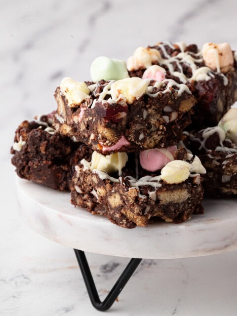A stack of delectable rocky road bars sits on a marble stand, inspired by a classic rocky road recipe. These treats are made of chocolate, nuts, and colourful marshmallows, drizzled with white chocolate. The background is softly blurred against a light marble surface.
