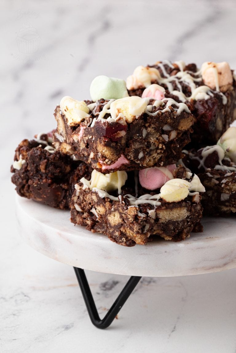 A stack of delectable rocky road bars sits on a marble stand, inspired by a classic rocky road recipe. These treats are made of chocolate, nuts, and colourful marshmallows, drizzled with white chocolate. The background is softly blurred against a light marble surface.